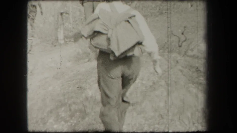 The woman continues down a rough path with a large bundle on her back in black-and-white 1938 home-movie footage.