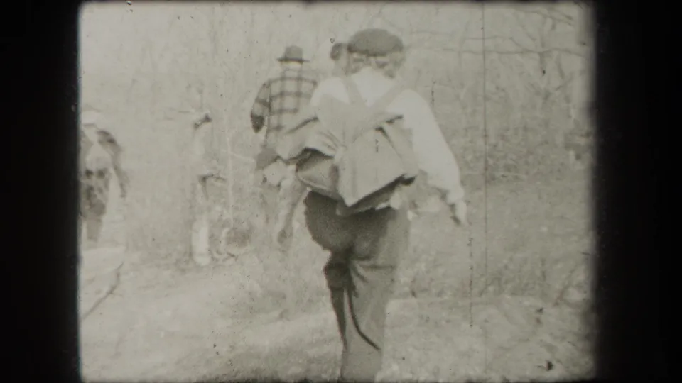 Two people walk away along a wooded path, with the nearer woman carrying a pack and another person ahead in a brimmed hat.