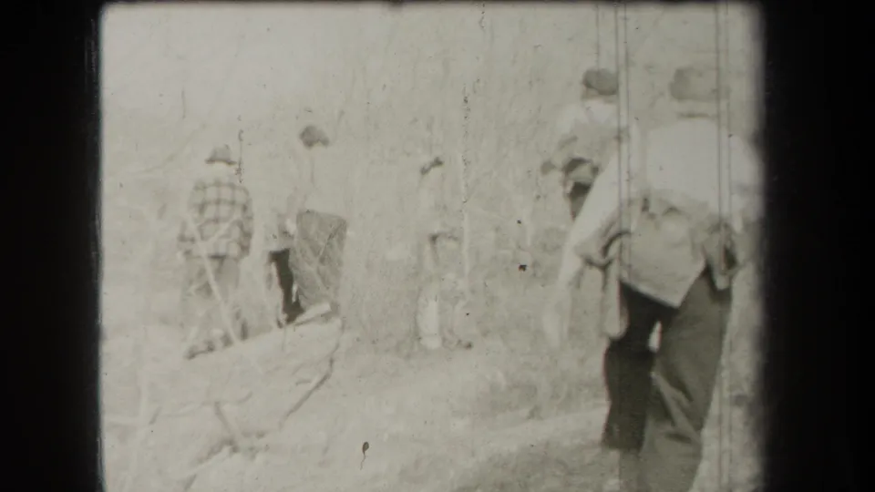 Rear-facing 8mm view of hikers in hats and workwear moving through a wooded New York trail in 1938.