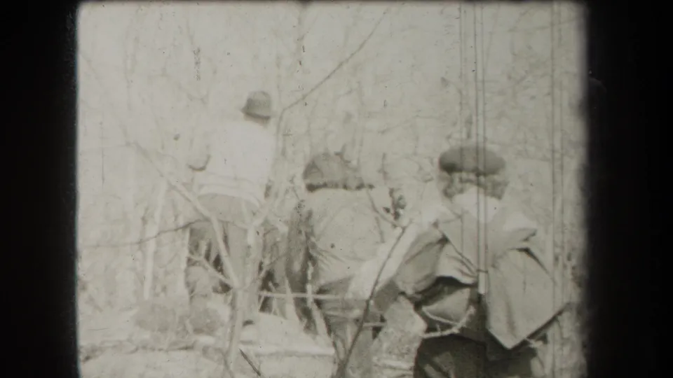A soft 1938 New York home-movie frame shows hikers from behind amid thin trees, brush, and a rough trail edge.