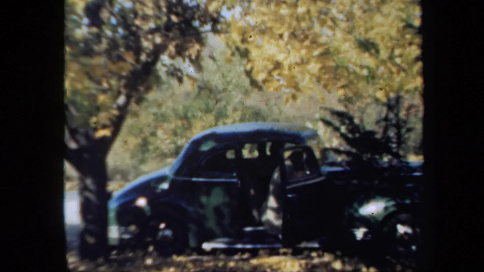 1939 New York home-movie frame with a person in period clothing emerging from a dark sedan under autumn trees.