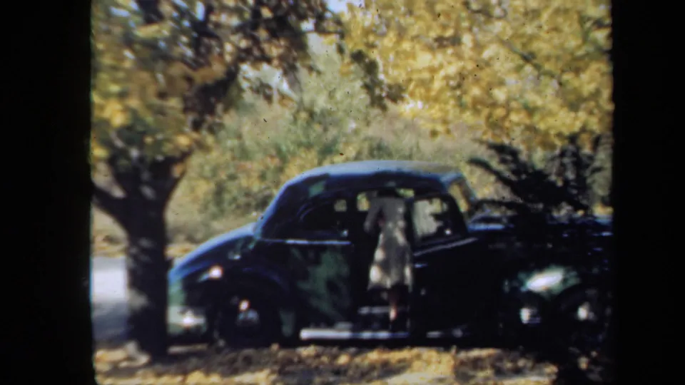 In 1939 New York home-movie footage, a woman stands at the open door of a dark 1930s automobile beneath yellow autumn trees.