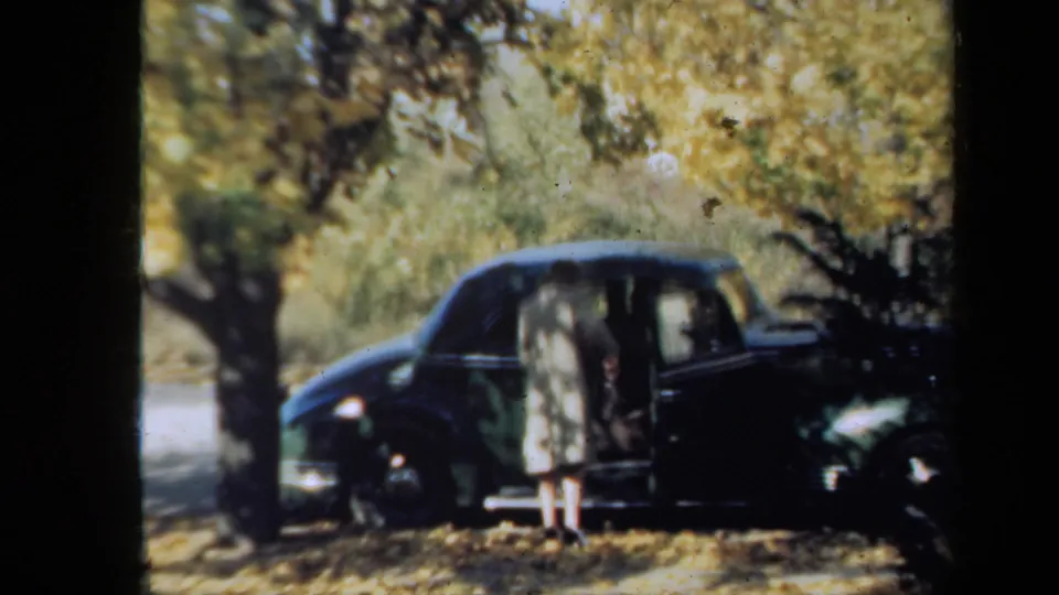 The woman has stepped down beside the dark 1930s car, with autumn leaves on the ground and trees behind her in supplied New York footage.