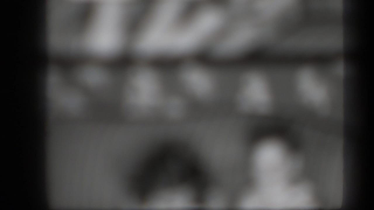 Blurred grayscale frame with two indistinct human figures behind dark film borders.