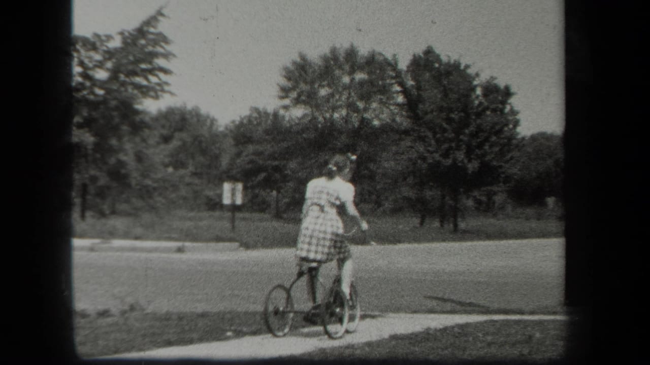 Girl on a small tricycle near a sidewalk and road with trees behind her.