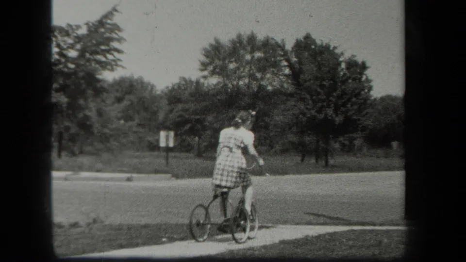 In 1939 New York home-movie footage, a girl in a light dress rides a small tricycle beside a quiet road and trees.