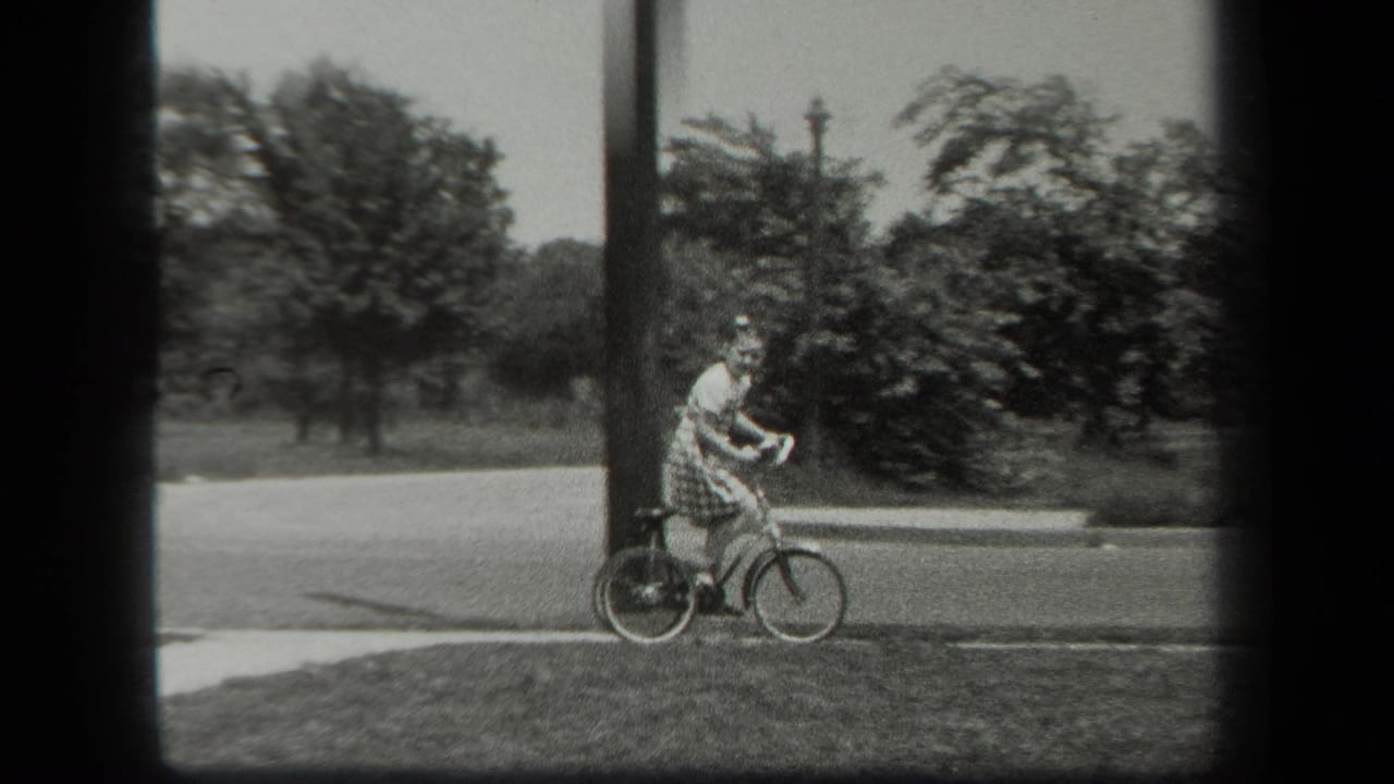 A girl on a small tricycle moves along a sidewalk edge in 1939 New York, passing a utility pole and roadside greenery.