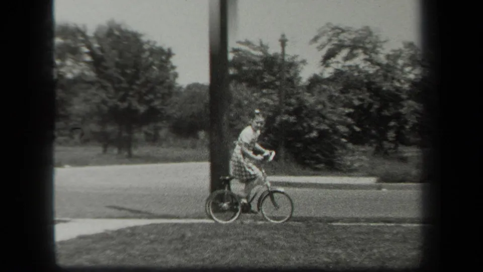 A girl on a small tricycle moves along a sidewalk edge in 1939 New York, passing a utility pole and roadside greenery.