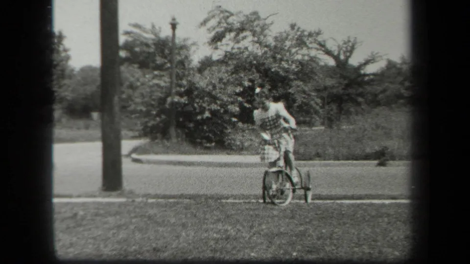 The girl appears to turn or steady her tricycle near the curb, framed by a utility pole, shrubbery, grass, and a quiet road.
