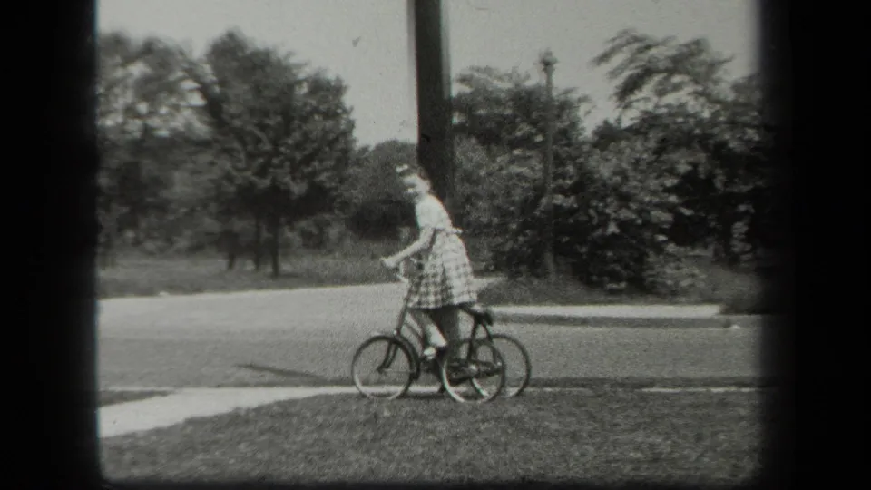 A girl in a light dress is seen with her small tricycle along a grassy sidewalk edge in the supported 1939 New York setting.