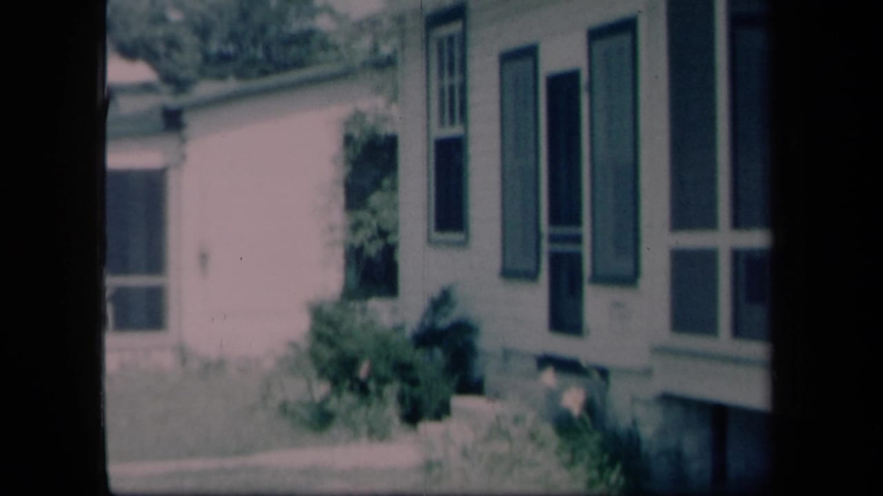 1958 New York frame of a white residential exterior with dark shutters, shrubs and a grass lawn.