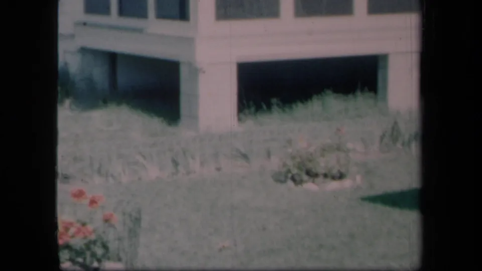 1958 New York 8mm frame showing a quiet house exterior with grass, flowers, windows, and part of a screened porch.