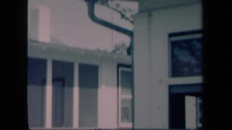 1958 New York 8mm frame showing the side of a house with porch supports, roofline, gutter, and adjacent windows.