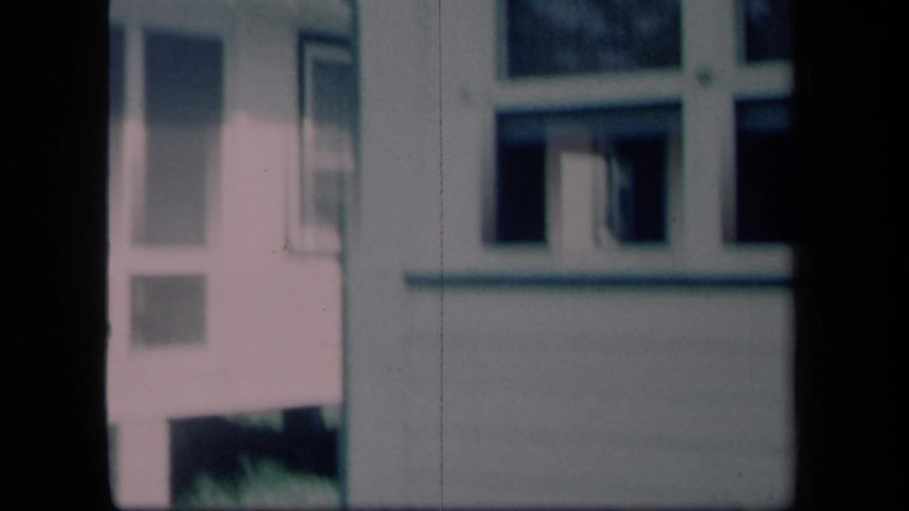 1958 New York 8mm frame showing a white residential exterior with porch structure, windows, and a dark railing or post in front.