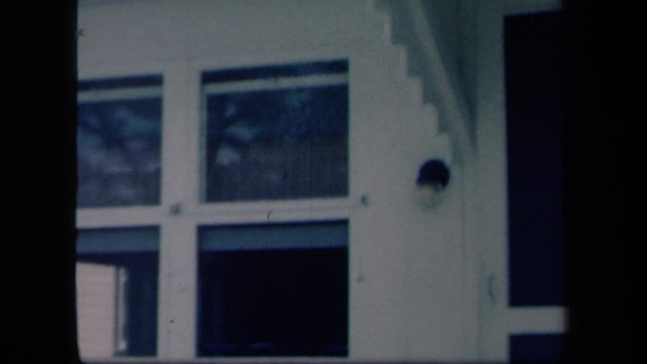 1958 New York home-movie frame of a white house facade with large windows, a dark porch opening, and vertical porch framing.