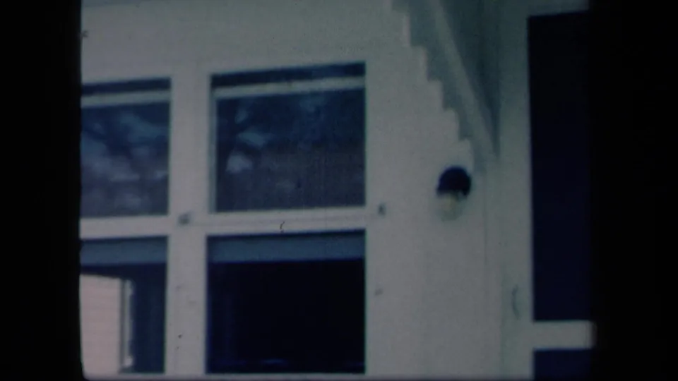 1958 New York home-movie frame of a white house facade with large windows, a dark porch opening, and vertical porch framing.