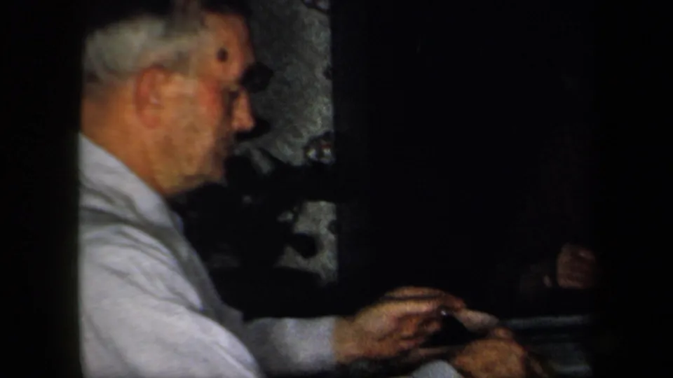 1958 New York home-movie frame showing an older man in profile at a dimly lit dining table inside a private interior.