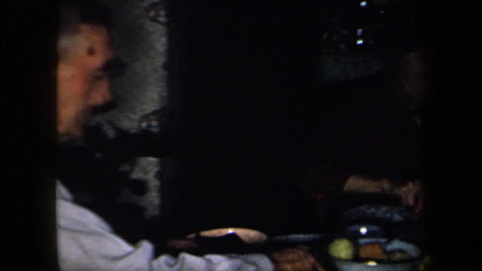1958 New York home-movie frame showing a low-light family meal, with an older man at left and a plate of food on the table.