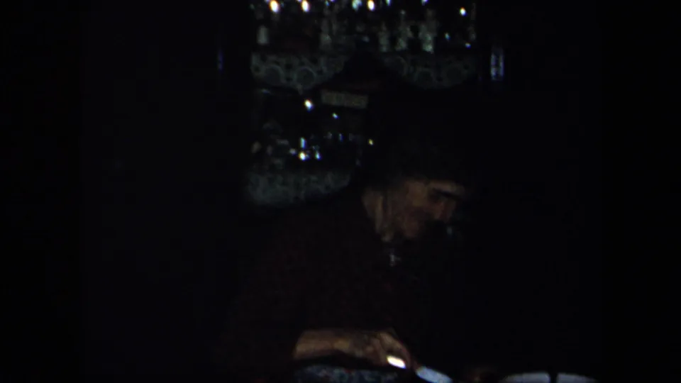 1958 New York home-movie frame showing a diner eating at a shadowed table, with reflective glassware or a chandelier above.