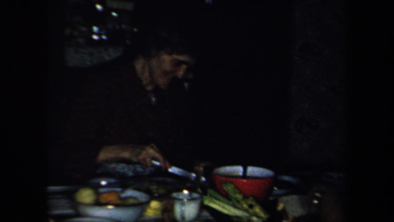 Adults seated at a dim dining table with dishes, vegetables, and a red bowl.