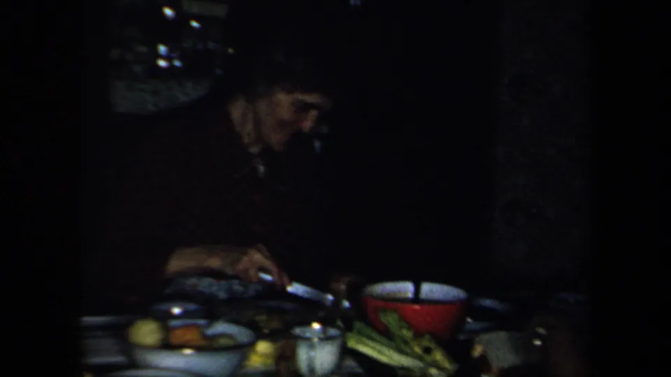 1958 New York home-movie frame of adults gathered at a dim indoor table with food, dishes, vegetables, and a red bowl.