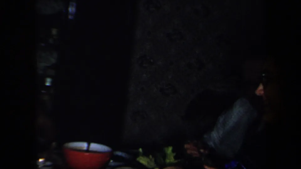 Low-light 1958 New York home-movie frame of a person seated beside a dining table with a red bowl and vegetables.