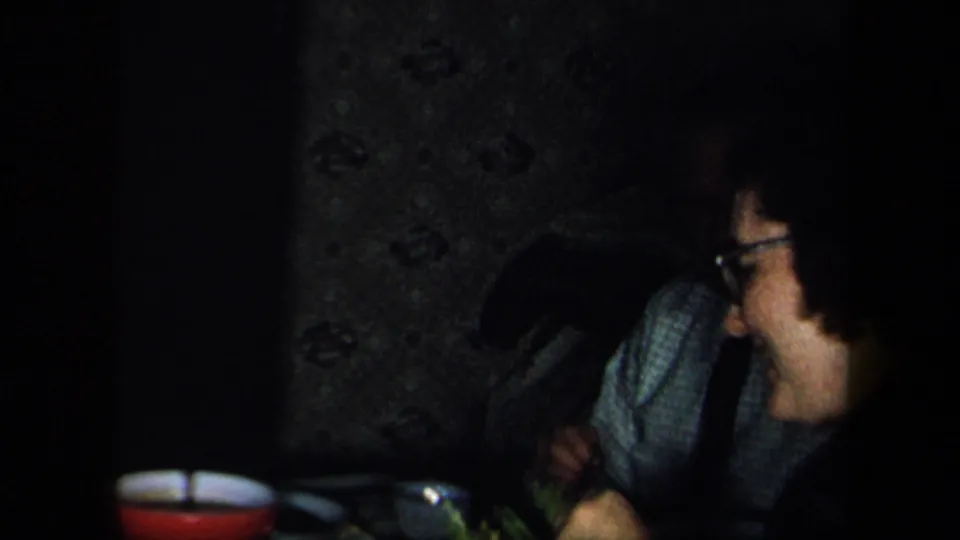 1958 New York home-movie frame of a person wearing glasses smiling beside a dim dining table with food and a red bowl.