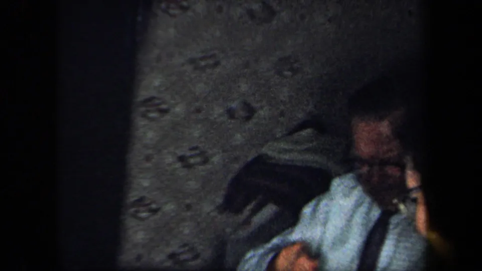 1958 New York home-movie frame showing a man in a shirt and tie seated near a table against a textured wall.