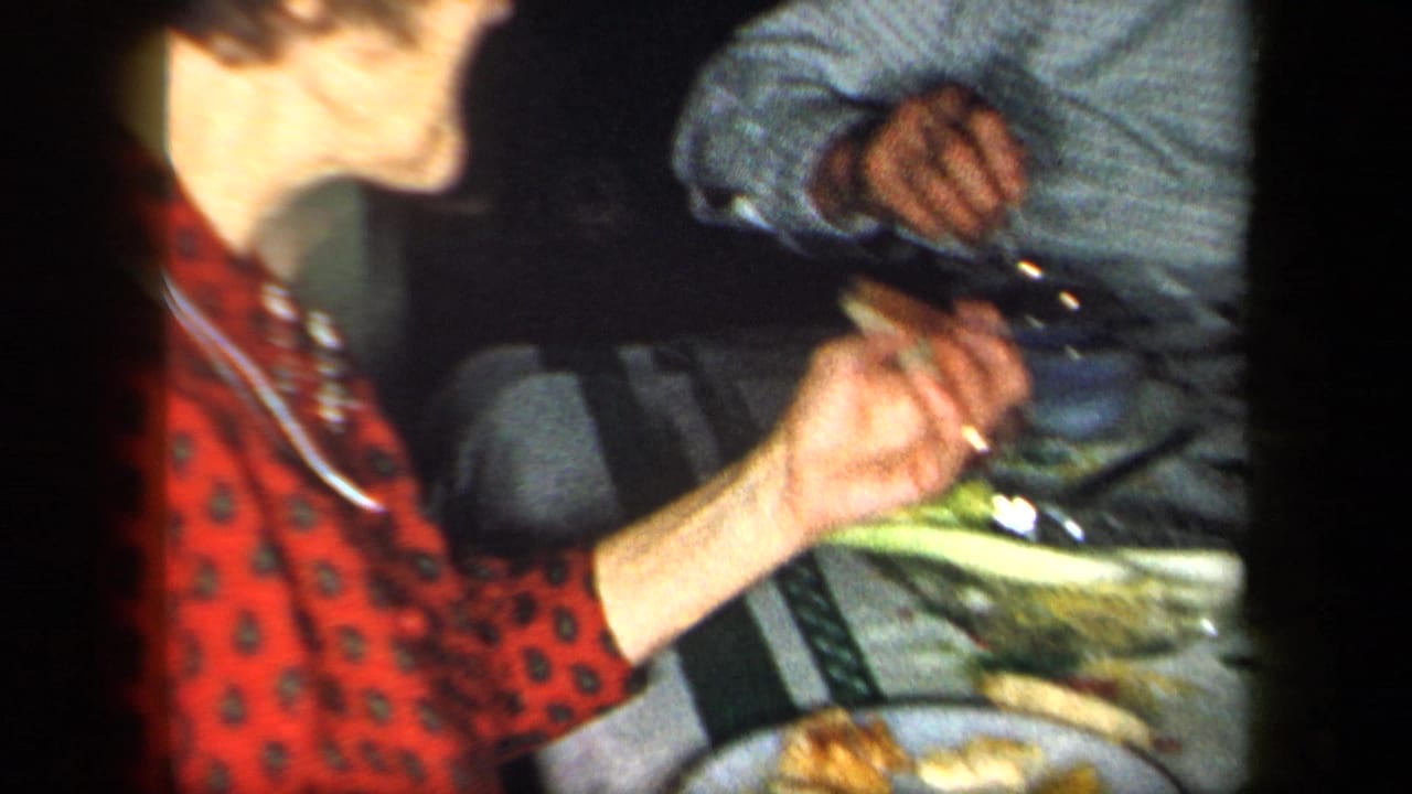 1958 New York home-movie frame of a woman in a red patterned blouse at a table with plates, food, and another seated adult nearby.