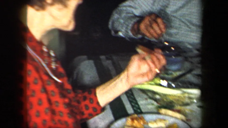 1958 New York home-movie frame of a woman in a red patterned blouse at a table with plates, food, and another seated adult nearby.