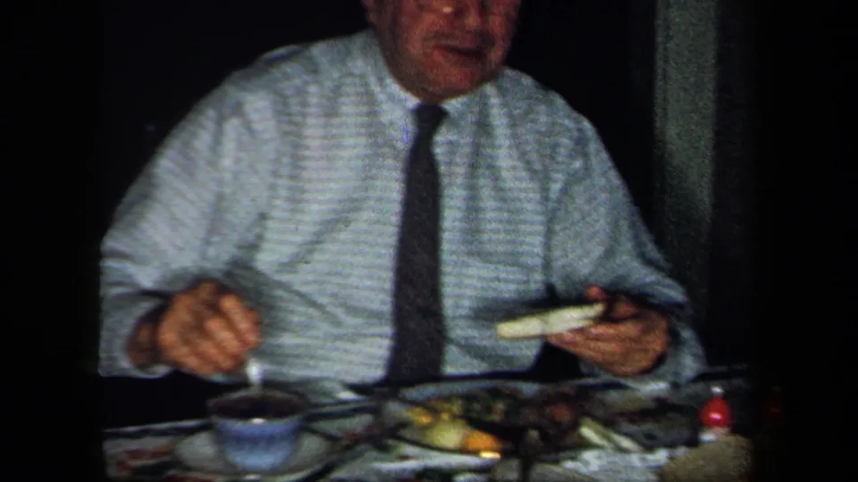 1958 New York home-movie frame showing an older man in a striped shirt and tie seated with a cup, plate, and food at the dinner table.