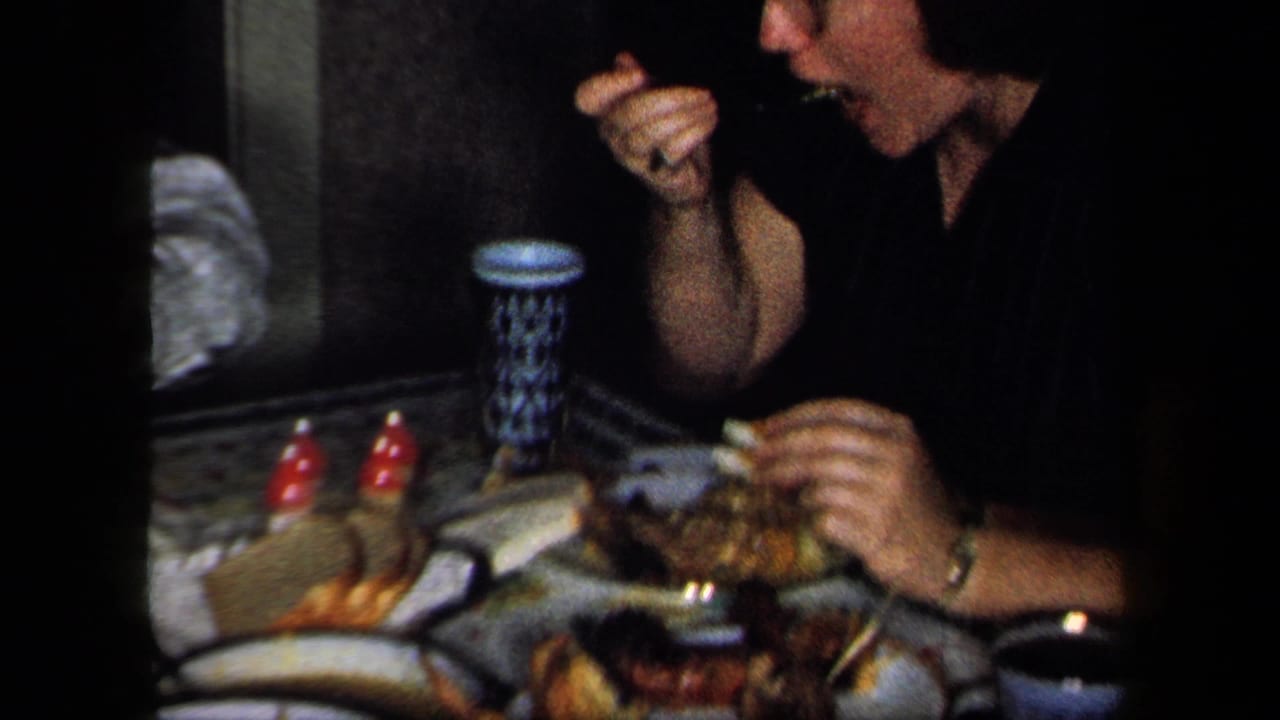 1958 New York home-movie frame showing a woman eating at a crowded dinner table with dishes, cups, and tableware.