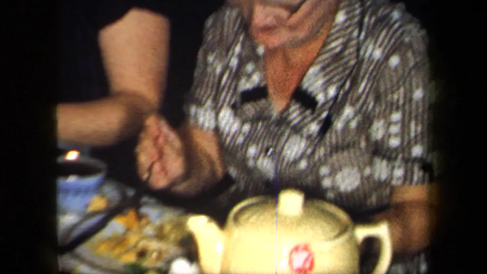 1958 New York home-movie still of an older woman at the dinner table with a yellow teapot, cups, and plates nearby.