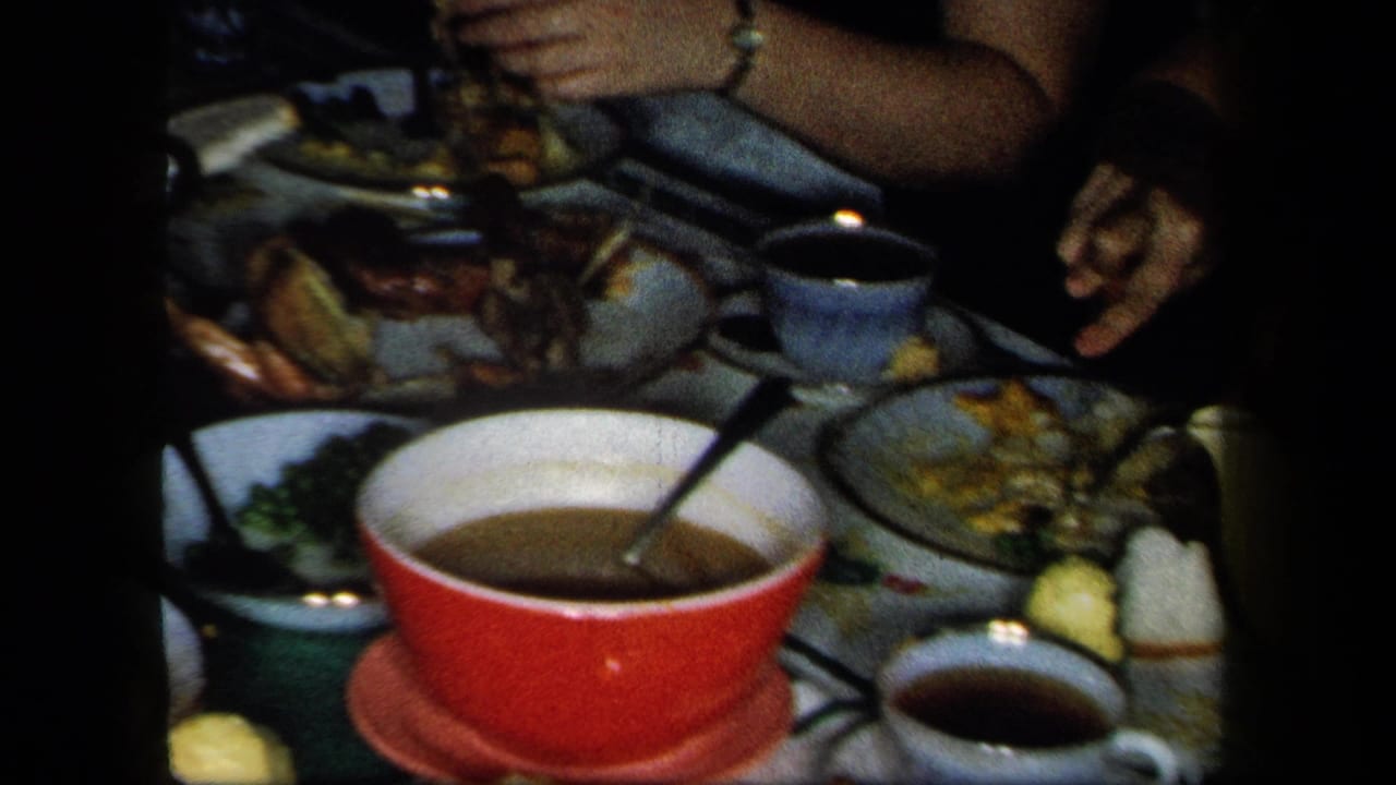 1958 New York home-movie frame of a dim dining table with a red bowl, teacups, plates of food, and diners' hands reaching in.