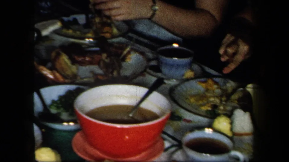 1958 New York home-movie frame of a dim dining table with a red bowl, teacups, plates of food, and diners' hands reaching in.