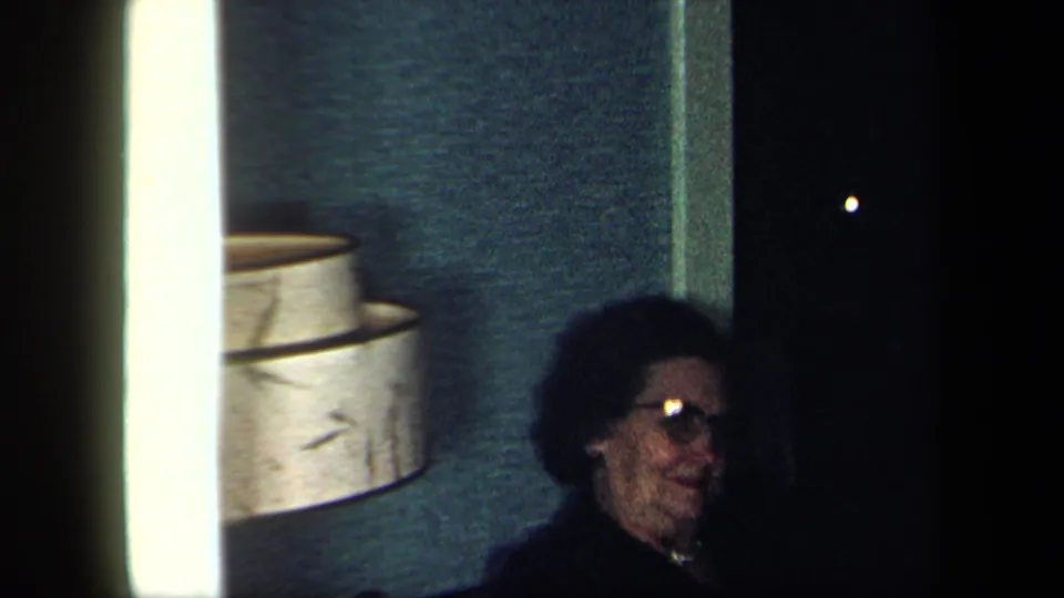 1958 New York home-movie frame of a man with glasses seated in a dim private dining-room interior near a wall lamp.