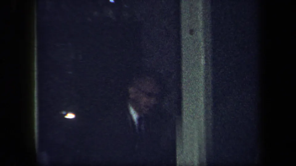 1958 New York home-movie frame with a dimly lit man partly visible near a wall or corner inside the private dining-room setting.