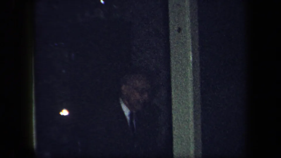 In a dim 1958 New York home-movie frame, a man in a dark suit and tie stands partly obscured near an interior doorway.