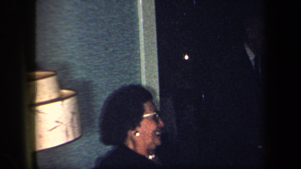 A woman with glasses and dark curly hair smiles in profile beside a table lamp in a dim New York interior from 1958.