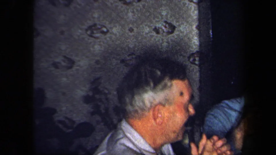 A man in a light shirt appears in profile against patterned wallpaper, with another figure partly visible in the low-lit 1958 interior.