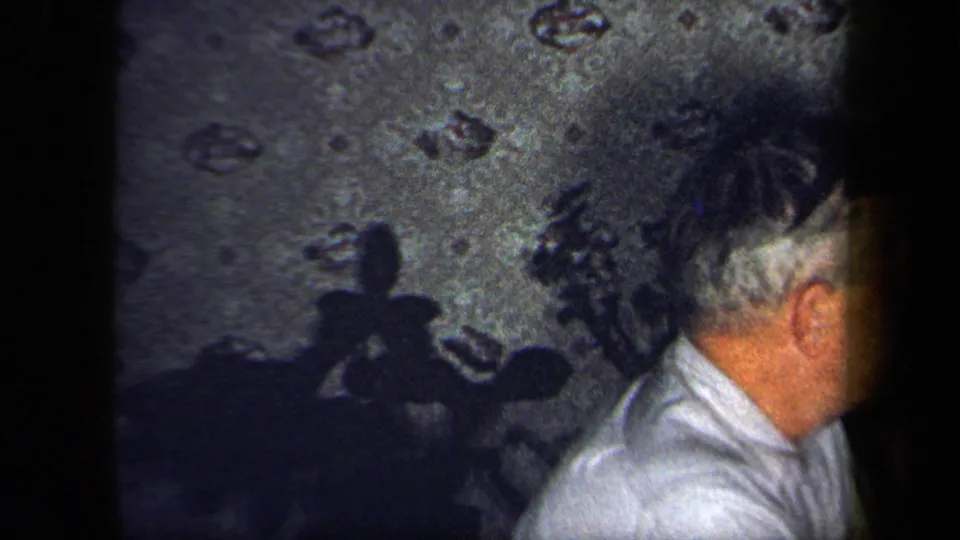 A gray-haired man in a light shirt moves across the frame as his shadow falls on patterned wallpaper in the 1958 New York interior.