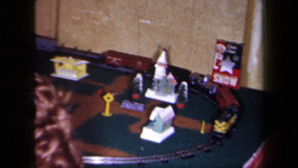 1958 New York home-movie frame showing a tabletop train village, small figures, curved track, and a partially readable holiday poster indoors.