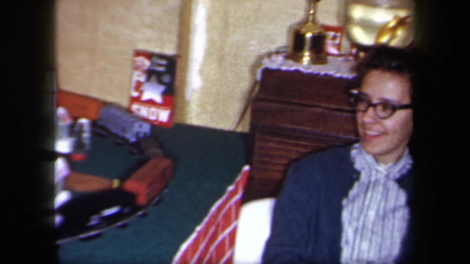 1958 New York home-movie frame of a smiling woman in glasses near a Christmas train display, dresser, lamp, and holiday poster.