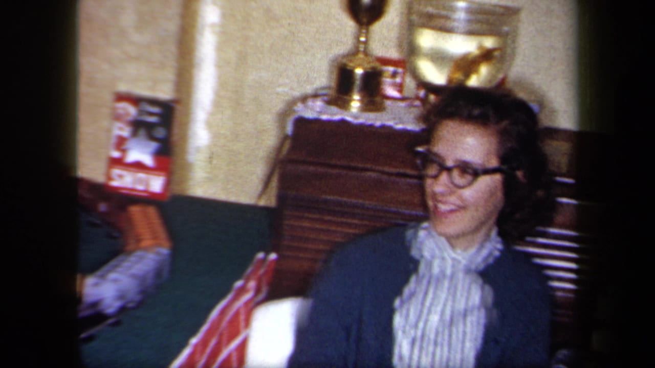 In a 1958 New York home-movie frame, a woman in glasses, a striped blouse, and cardigan smiles beside wooden furniture and display items.