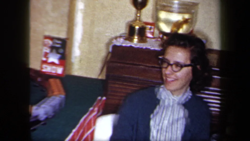 In a 1958 New York home-movie frame, a woman in glasses, a striped blouse, and cardigan smiles beside wooden furniture and display items.