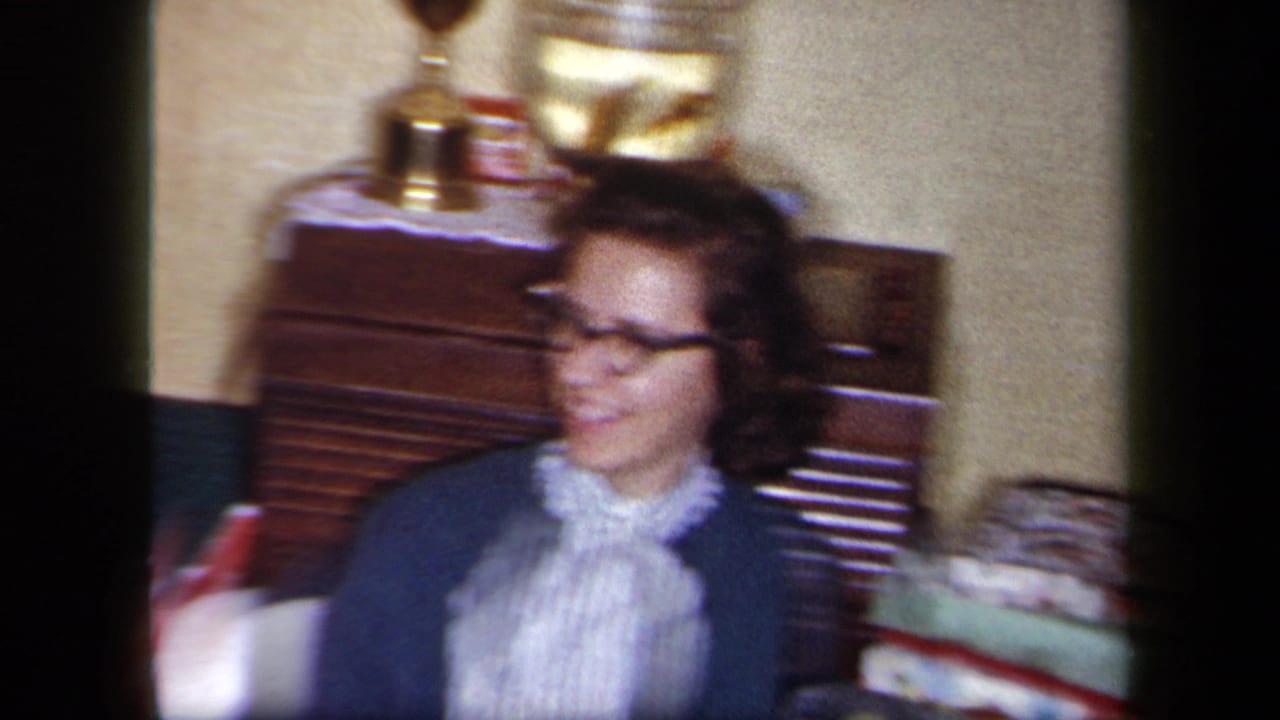 A 1958 New York home-movie frame shows a woman in glasses turning while seated near wooden furniture, display objects, and household items.