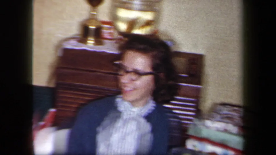 A 1958 New York home-movie frame shows a woman in glasses turning while seated near wooden furniture, display objects, and household items.