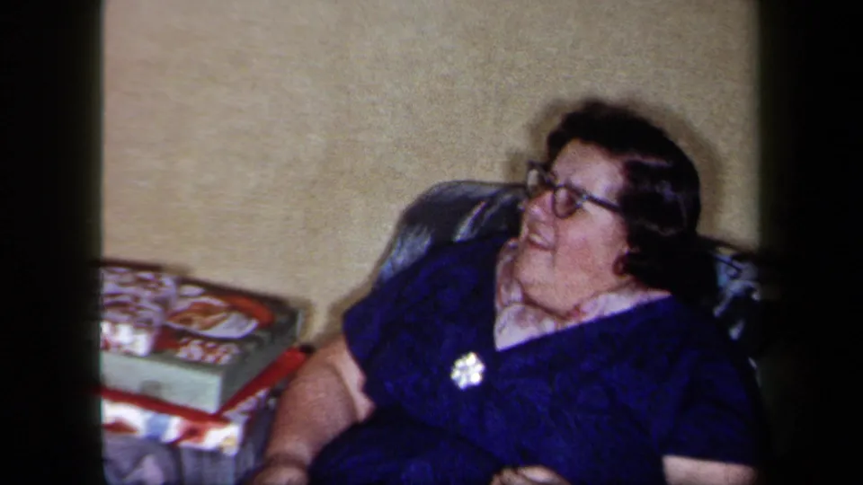 In this 1958 New York home-movie frame, a woman wearing glasses and a dark blue dress sits beside boxed tabletop games in a living room.