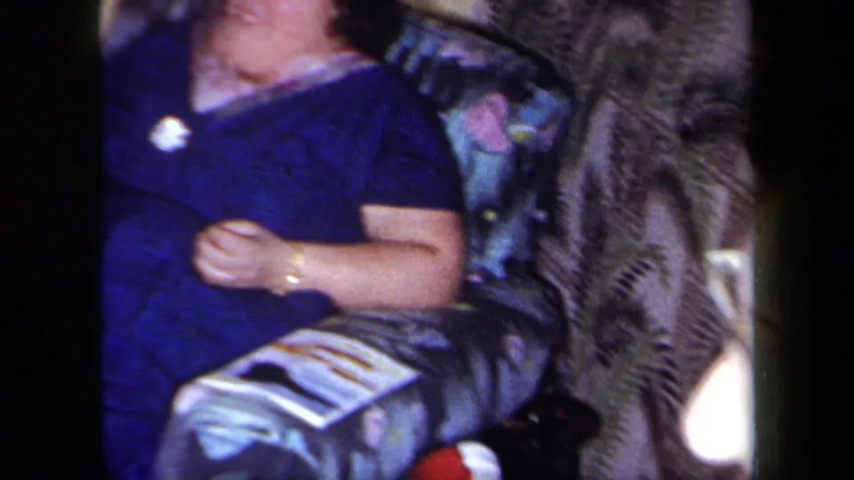 A 1958 New York home-movie frame shows a woman in a blue dress seated in a patterned chair, with boxed games and indoor decor nearby.