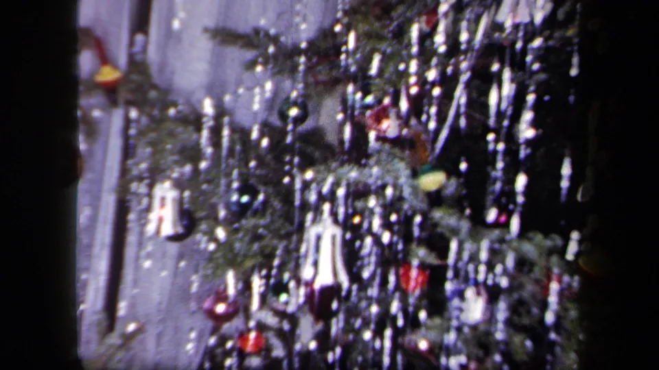 1958 New York 8mm frame showing a close domestic view of a Christmas tree with tinsel, ornaments, and a pale interior wall behind it.